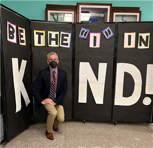 Superintendent Tom Rogers standing in front of sign that reads "Be the 'I' in Kind"